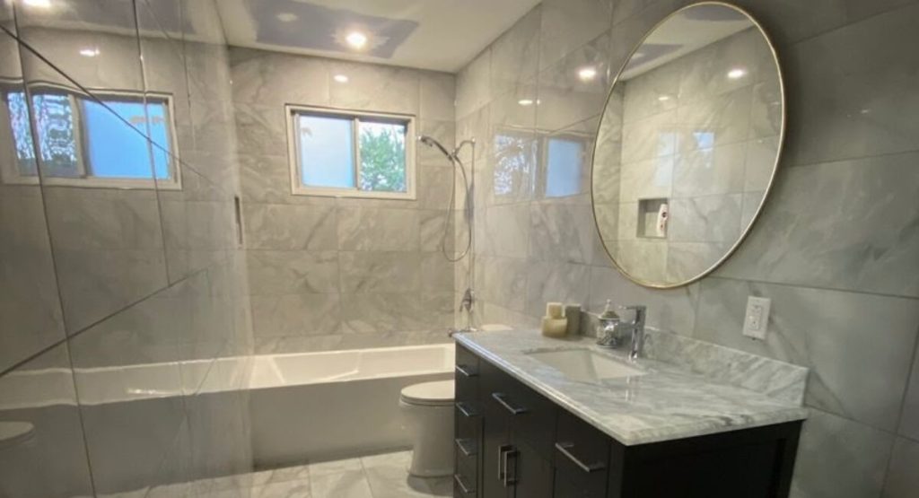 Remodeled bathroom with marble-look porcelain tile, a tiled tub-shower and a round mirror over a dark vanity