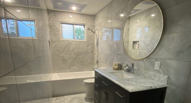 Remodeled bathroom with marble-look porcelain tile, a tiled tub-shower and a round mirror over a dark vanity
